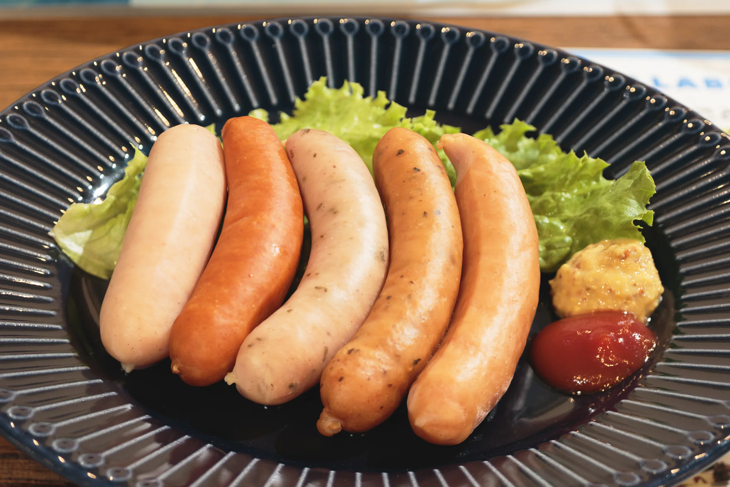 黒い皿に盛り付けられた5本のソーセージとレタス，マスタード，ケチャップ