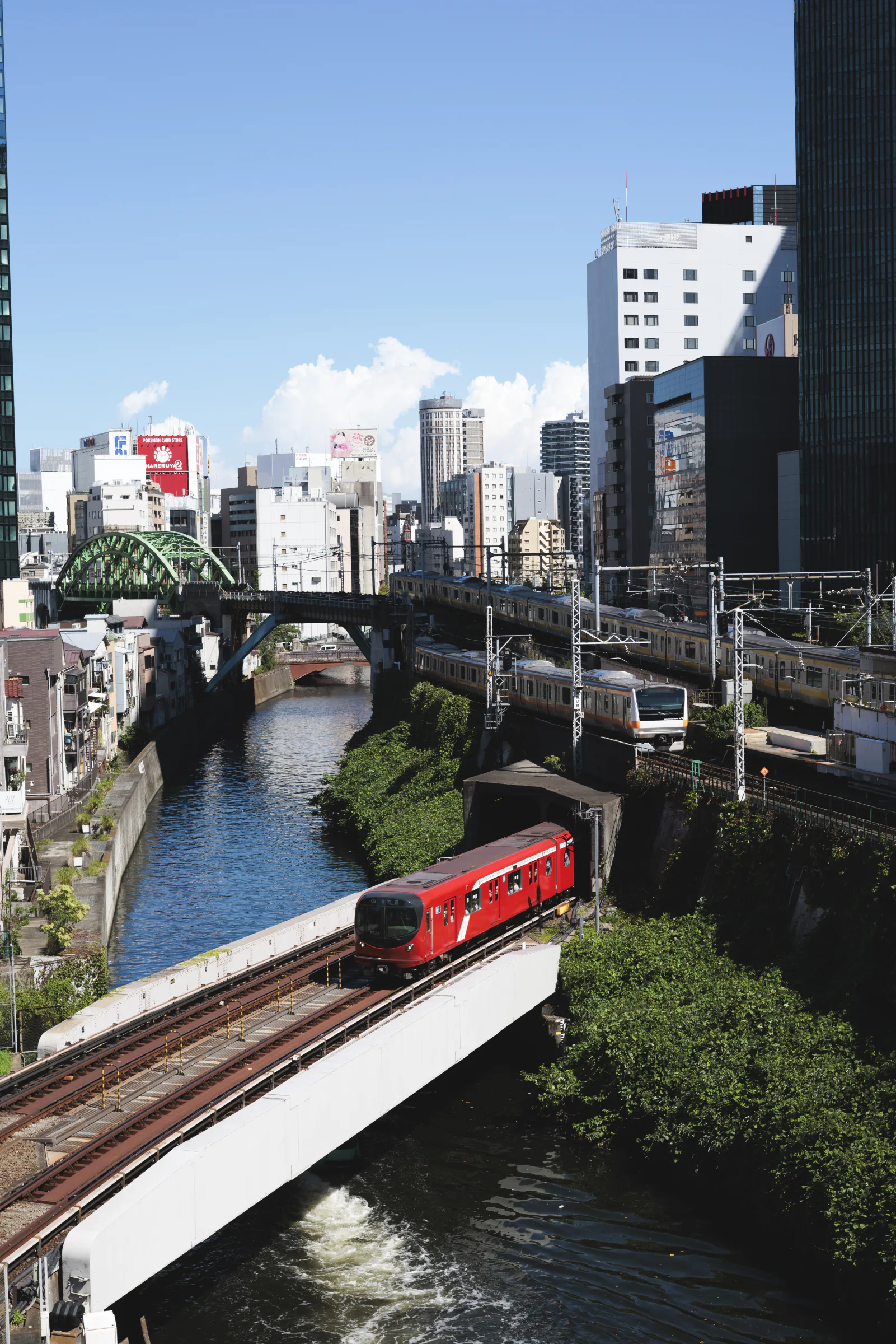 青空の下，川沿いを走る赤と銀色の列車を高所から捉えた都市風景